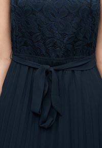 Φόρεμα σε ναυτικό μπλε με μπούστο από δαντέλα με λουλούδια, πλισέ φούστα και δεμένη υφασμάτινη ζώνη στη μέση.