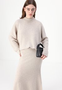 Beige gestrickter Pullover mit hohem Kragen und lockeren Ärmeln, kombiniert mit einem passenden taillierten Rock. Das Modell hält eine kleine schwarze Handtasche.