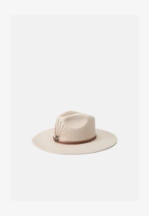 Sombrero de fieltro beige de ala ancha que presenta una banda decorativa de cuero marrón y acentos metálicos con detalles alargados en el lado.