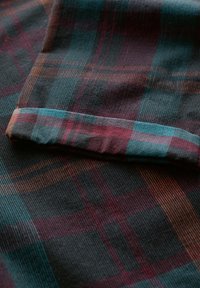 Tessuto a quadri con sfondo scuro, caratterizzato da linee in teal, borgogna e marrone. Superficie testurizzata con righe verticali e bordo piegato.