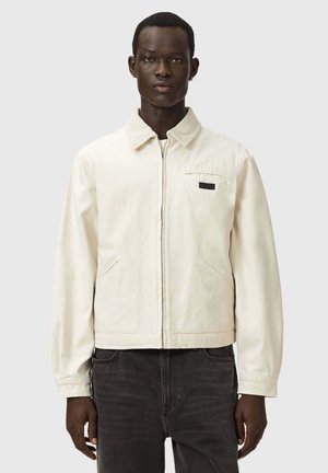 Mand iført en lys beige lynlåsjakke med krave og mørke jeans, stående foran en ensfarvet lysegrå baggrund.