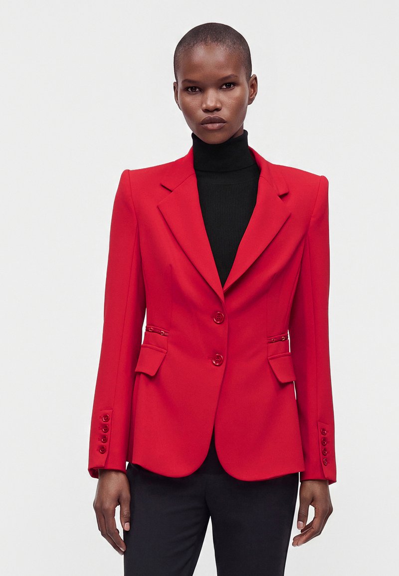 Femme portant un blazer rouge vif ajusté avec des épaules structurées par-dessus un col roulé noir et un pantalon noir, sur un fond gris uni.