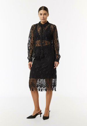 Black lace two-piece set featuring a long-sleeve shirt and knee-length skirt, floral patterns, and a scalloped hem. Accessories include black heels.
