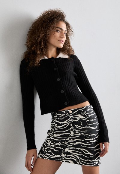 Μαύρο ριμπ πλεκτό κοντό πουλόβερ με λευκό γιακά και έξι κουμπιά, σε συνδυασμό με μαύρη και άσπρη τζιν φούστα με τυπώμα ζέβρας που διαθέτει μπροστινά κουμπιά.