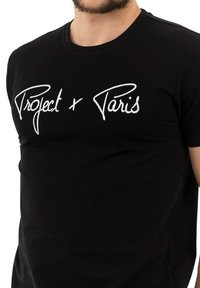 Camiseta de algodón negra con texto en cursiva blanca "Project x Paris" en el frente. Mangas cortas y cuello redondo con un diseño ajustado.