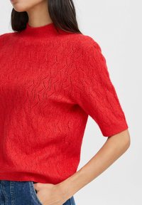 Roter gestrickter Pullover mit kurzen Ärmeln, der ein strukturiertes Wellenmuster und einen gerippten hohen Kragen aufweist. Glatte Oberfläche, lässige Passform.