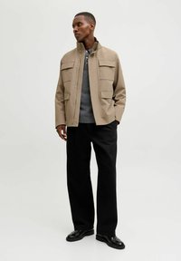 Bežna utilitarna jakna z visokim ovratnikom, z dvema sprednjima žepoma, nošena čez sivo pulover in črne široke hlače, v kombinaciji s črnimi čevlji.