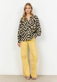 Halenka s dlouhými rukávy v krémové barvě s černým zigzag vzorem, v kombinaci se žlutými kalhotami s širokými nohavicemi. Obsahuje knoflíkovou légu a volný střih.