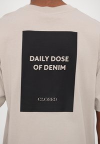 T-shirt en coton beige avec un graphique rectangulaire noir au dos. Le texte indique "DOSE QUOTIDIENNE DE DENIM" et "CLOSE" en blanc. Manches courtes.