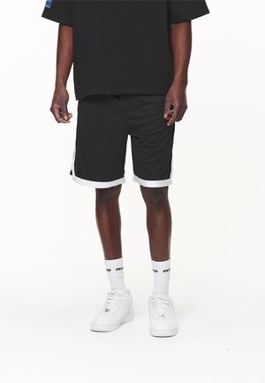 Mann trägt schwarze Sportshorts mit weißem Besatz, weiße Socken mit schwarzem Text und weiße Turnschuhe, steht auf weißem Hintergrund.