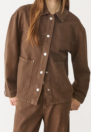 Femme portant une veste marron boutonnée avec de grandes poches avant et un pantalon marron assorti, debout devant un fond clair.