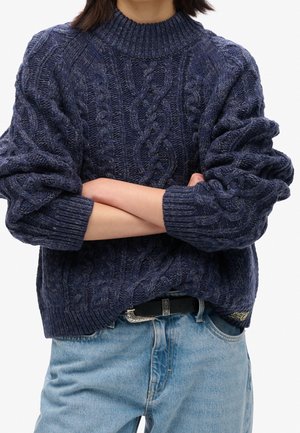 Osoba ubrana w granatowy sweter z warkoczowym splotem, z podwiniętymi rękawami, skrzyżowanymi ramionami, jasnoniebieskie dżinsy i czarny pasek z srebrną klamrą.