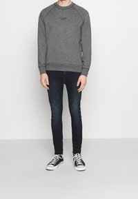 Sudadera gris de cuello redondo con mangas raglán que presenta un logo sutil, combinada con jeans oscuros y zapatillas en blanco y negro.