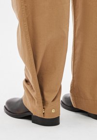 Brązowe bawełniane spodnie o luźnym kroju, zdobione złotymi detalami przy mankietach, zestawione z czarnymi botkami za kostkę.