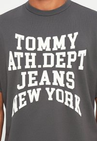 Šedé bavlnené tričko s výrazným bielym textom: "TOMMY ATH.DEPARTMENT JEANS NEW YORK." Klasický strih s okrúhlym výstrihom a krátkymi rukávmi.