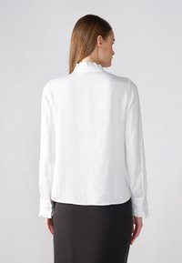 Blouse blanche à manches longues avec un col volanté et des poignets boutonnés, présentant un tissu doux et texturé ainsi qu'une silouhette à coupe décontractée.