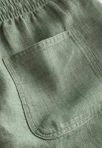 Gros plan d’un pantalon en tissu vert clair avec une taille élastique et une poche carrée cousue visible au centre.