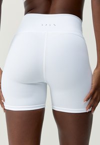 Shorts de sport blancs à taille haute, avec une coupe ajustée, dotés de coutures plates et d'une texture lisse. Logo imprimé sur la ceinture.