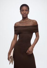 Γυναίκα με εφαρμοστή καφέ κοντή off-shoulder φόρεμα κρατώντας ένα μπεζ τσαντάκι, φορώντας χρυσά κοσμήματα, πάνω σε απλό λευκό φόντο.