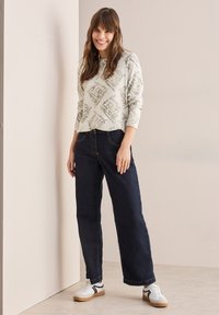 Hellgrauer Pullover mit Rautenmuster; lockere dunkle Jeans; weiße Sneakers mit schwarzen Akzenten; glatte Texturen; entspannte Passform.