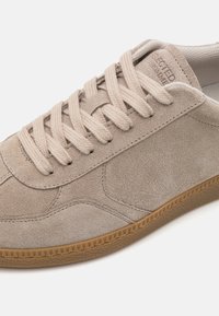 Beige mockasinsko med rund tå, texturerad yta och matchande snörning. Gummisula med mönstrad kant för bra grepp. Minimalistisk design.