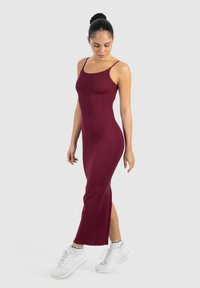 Mujer con un vestido ajustado sin mangas de color burdeos y zapatillas blancas, de pie y mirando ligeramente hacia abajo sobre un fondo liso y claro.