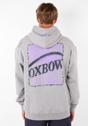 Sweat-shirt gris avec capuche, arborant un grand graphique violet et du texte noir au dos. Texture douce, coupe décontractée et poignets côtelés.