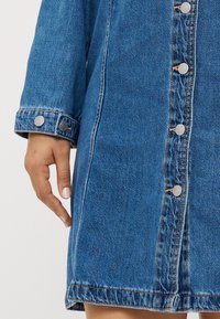 Denim jaka vidēji zilā krāsā ar sudrabainiem uzspiediet pogām, redzamām šuvēm un nedaudz saplēstu apakšmalu. Raksturīga brīvā piegriezuma dizainam un aproču detaļām.