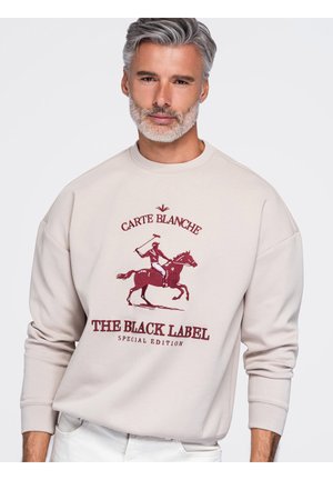Mężczyzna z siwymi włosami i brodą, ubrany w beżową bluzę z czerwoną grafiką gracza polo i tekstem "Carte Blanche The Black Label Special Edition."