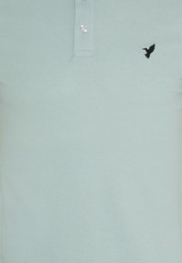 Polo vert clair avec deux boutons blancs et un petit logo d'oiseau noir brodé sur le côté gauche de la poitrine.