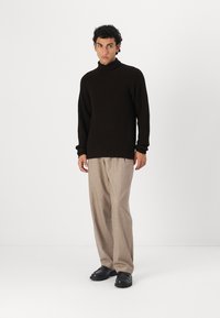 Zwarte geribbelde coltrui, losse pasvorm, met beige high-waisted broek en zwarte schoenen, gedragen door een model in een minimalistische setting.