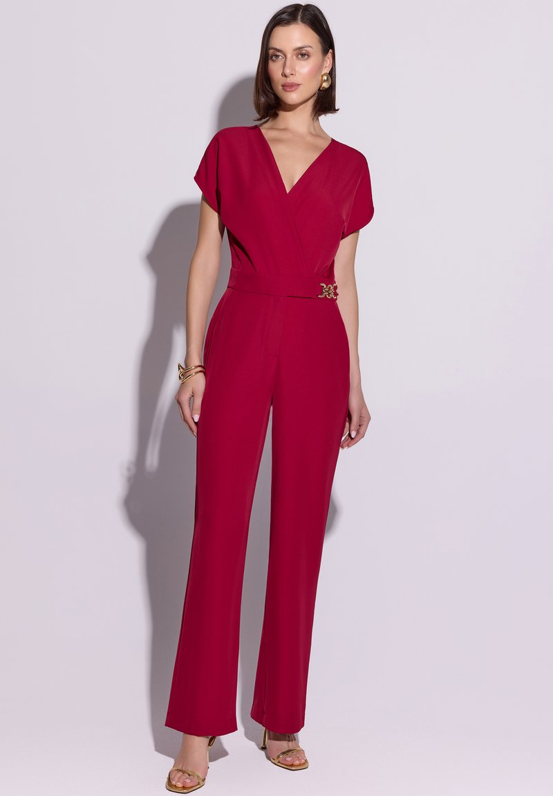 Kobieta stojąca, ubrana w głęboko czerwony kombinezon z dekoltem w serek, krótkimi rękawami, szerokimi nogawkami, złotymi kolczykami, bransoletkami i sandałami na obcasie.