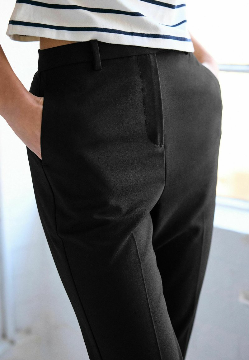 Personne portant un pantalon noir sur mesure avec les mains dans les poches et une chemise blanche à rayures horizontales noires.