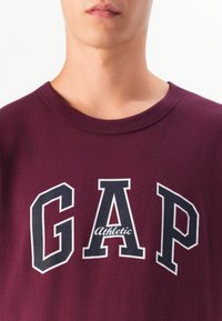 Oberkörper einer Person, die einen weinroten Rundhalsausschnitt-Sweatshirt trägt, auf dem große schwarze und weiße Buchstaben "GAP Athletic" auf der Vorderseite stehen.