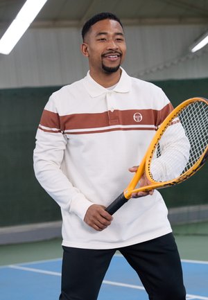 Valkoinen pitkähihainen polo, jossa on ruskea raita ja logo; pelaaja pitää käsissään oranssia tennismailaa sinisellä kentällä vihreän taustalla.
