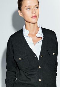 Cardigan nero con bottoni dorati, dotato di due tasche frontali. Abbinato a una camicia bianca con colletto, che presenta un design semplice e tessurizzato.