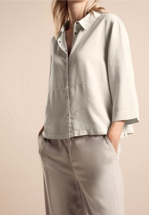 Hellgraues Hemd mit Knopfleiste, Kragen, kurzen Ärmeln und dezenten Nahtdetails, kombiniert mit glatten beigen Hosen.