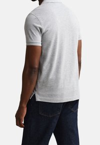 Homme portant un polo gris clair à manches courtes et un jean bleu foncé, debout dos à la caméra devant un fond uni.