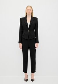 Completo nero su misura con giacca aderente caratterizzata da spalle slanciate e bottone dorato, abbinato a pantaloni slim-fit e tacchi neri a punta.