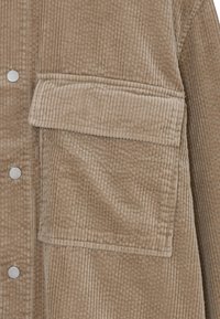 Veste en velours côtelé marron avec une grande poche poitrine, des boutons-pression en métal argenté et une texture à côtes verticales. Tissu de haute qualité avec une finition douce.