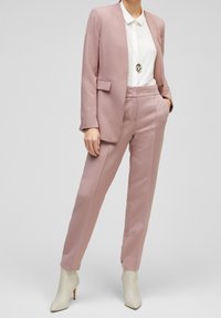 Ljust rosa kostymjacka med en enda ficka och matchande skräddarsydda byxor. Överdimensionerad krage på vitt blus under och beige högklackade stövletter.