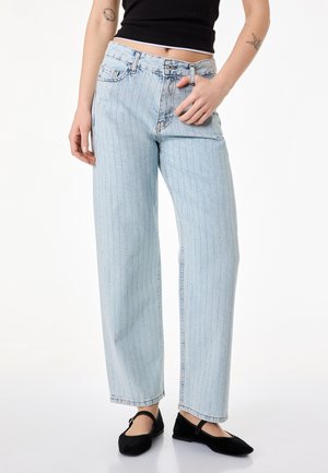 Kvinde iført lyseblå højtaljede jeans med lodrette prikstriber, sort ærmeløs top og sorte Mary Jane-flade sko.