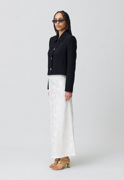 Chaqueta negra corta con botones dorados, combinada con una falda maxi blanca con flores y sandalias de tacón amarillas. El atuendo presenta un corte estructurado.