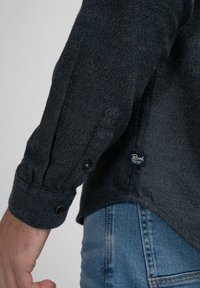 Donkerblauwe lange mouw shirt van gestructureerde stof, met knopenmanchetten en een klein logo-label aan de zijkant, gecombineerd met lichtblauwe denim jeans.