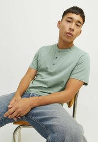 Camisa henley de manga corta verde claro con tres botones, combinada con unos vaqueros de mezclilla azul deslavado, llevada mientras se está sentado en una silla de madera.