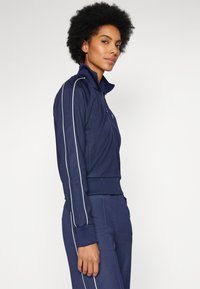 Giacca sportiva con zip di colore navy, con strisce bianche sui lati, polsini e vita a coste. Texture liscia e design aderente.