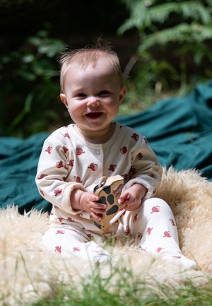 Helle cremefarbene Einteiler mit roten Affenmuster, langen Ärmeln und Druckknöpfen. Kind hält ein hölzernes Giraffenspielzeug. Weicher Teppich aus Fell darunter.
