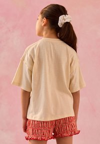 T-shirt beige oversize à manches courtes, associé à un short volanté à carreaux rouges et blancs. Le fond est une texture rose douce.