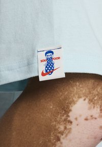 T-shirt en coton bleu clair avec une étiquette présentant un design graphique d'un personnage et le texte "SOLO FOOD", accompagné d'un logo rouge de Nike.