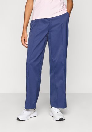 Granatowe spodnie o prostych nogawkach, wykonane z gładkiej tkaniny, z bocznymi kieszeniami i dopasowanymi szwami, zestawione z białymi butami sportowymi.